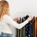 A woman in a white shirt browsing a clothing rack of shirts and sweaters while avoiding fall closet-swap mistakes.