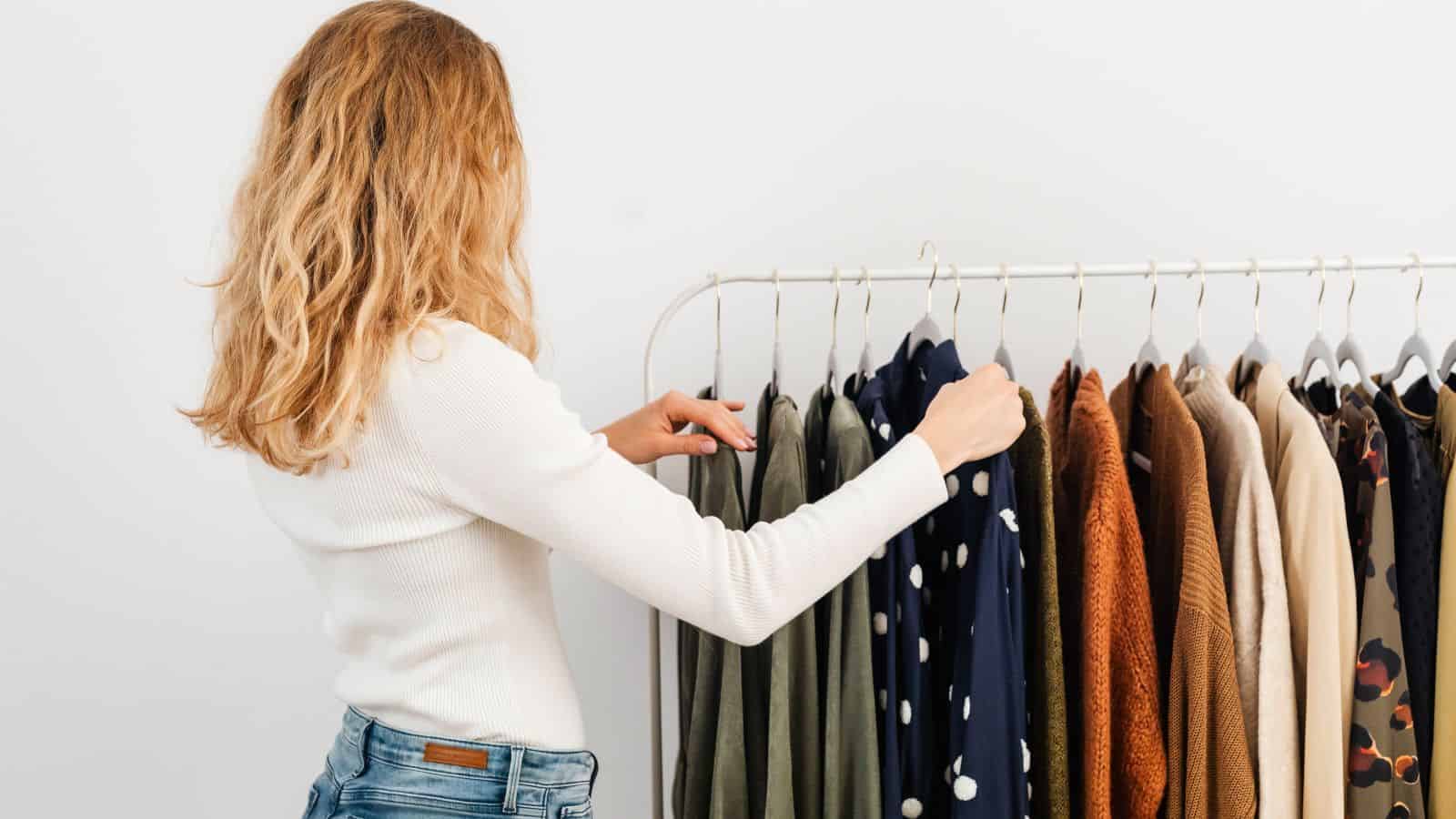 A woman in a white shirt browsing a clothing rack of shirts and sweaters while avoiding fall closet-swap mistakes.