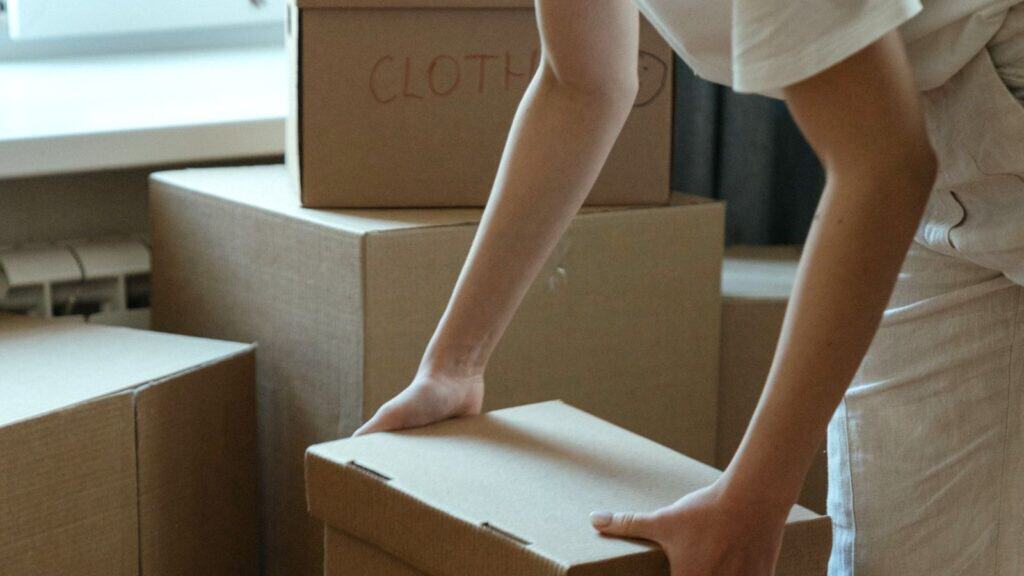 A person packs a cardboard box among others, including one labeled &ldquo;CLOTHES,&rdquo; while avoiding common fall closet-swap mistakes.