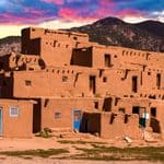 Multi-story adobe structure at Taos Pueblo against a mountain backdrop under a colorful sunset sky.