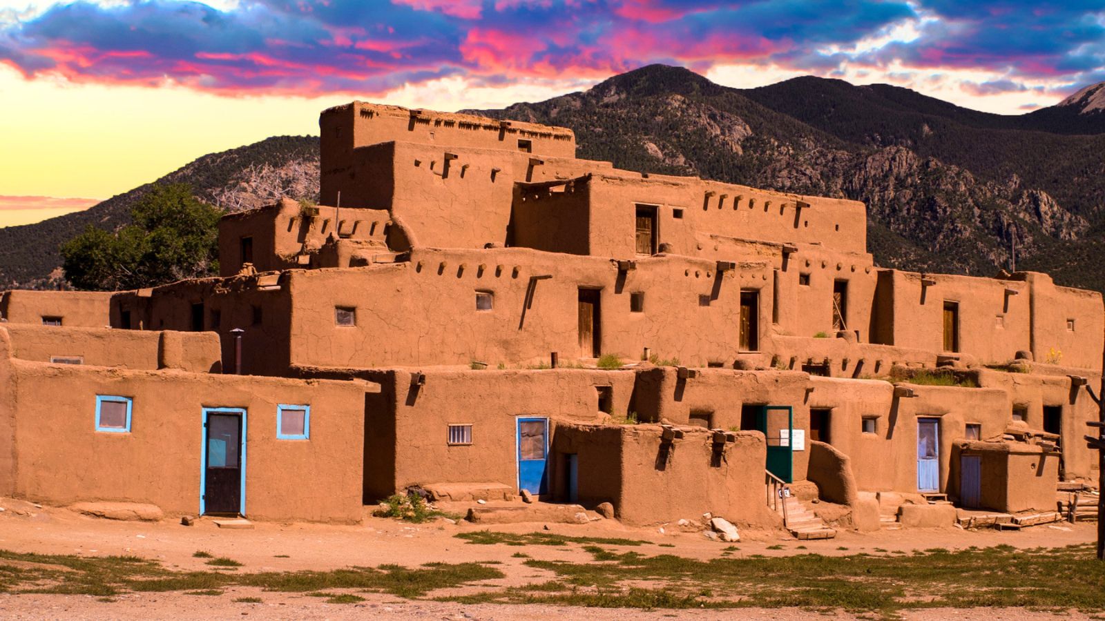 Multi-story adobe structure at Taos Pueblo against a mountain backdrop under a colorful sunset sky.