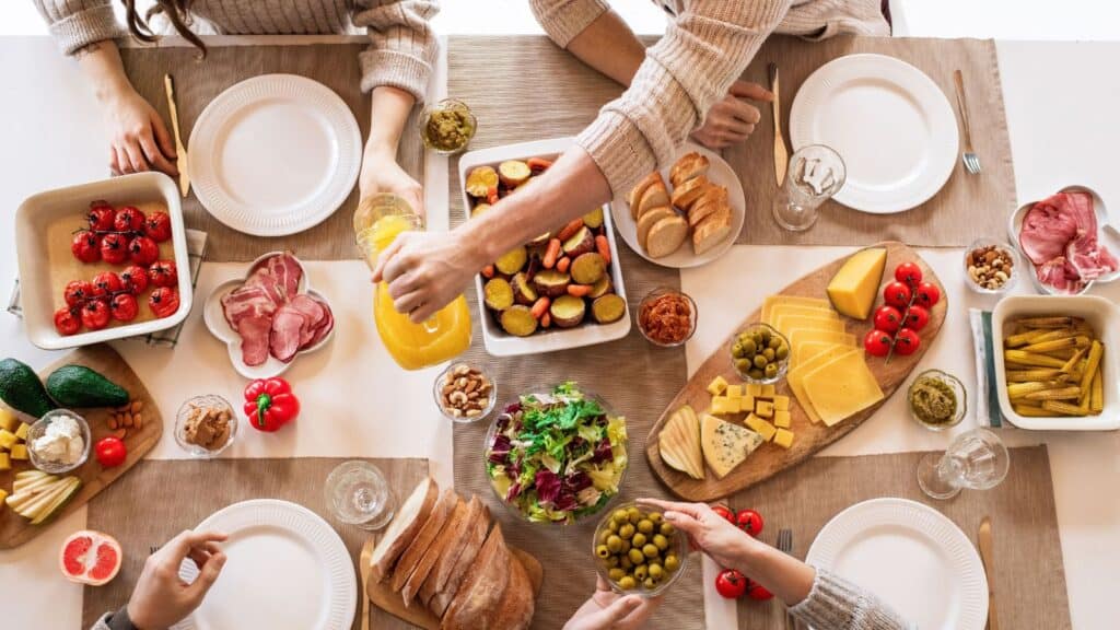 People gather around a table of World Food Day appetizers, including roasted vegetables, sliced meats, cheeses, and bread, as one person pours orange juice.