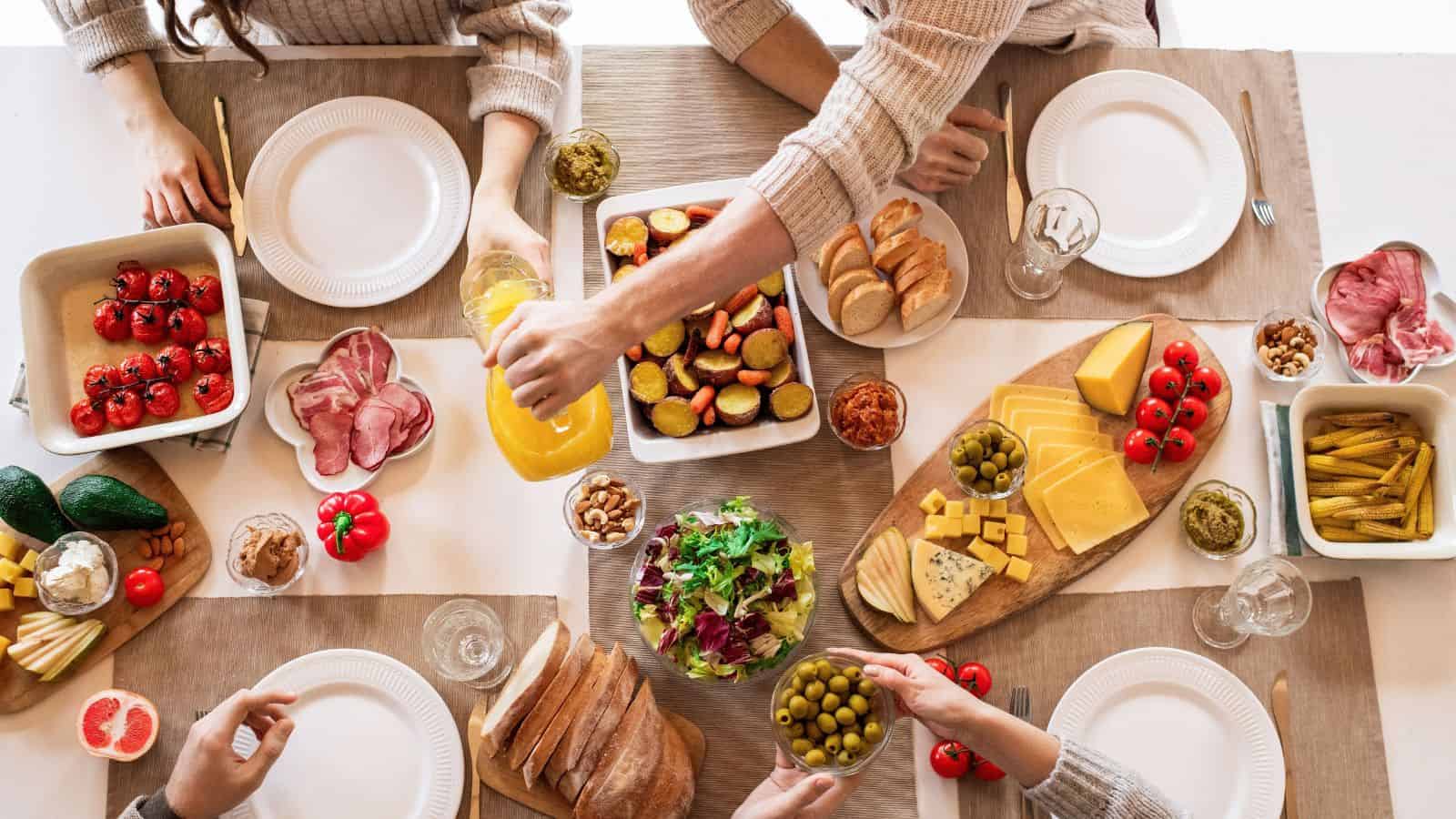 People gather around a table of World Food Day appetizers, including roasted vegetables, sliced meats, cheeses, and bread, as one person pours orange juice.