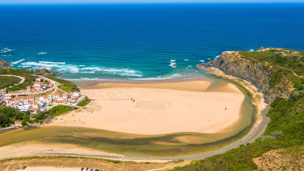Aerial view of a coastal beach with a small village, a river estuary flowing into the ocean, and surrounding cliffs.