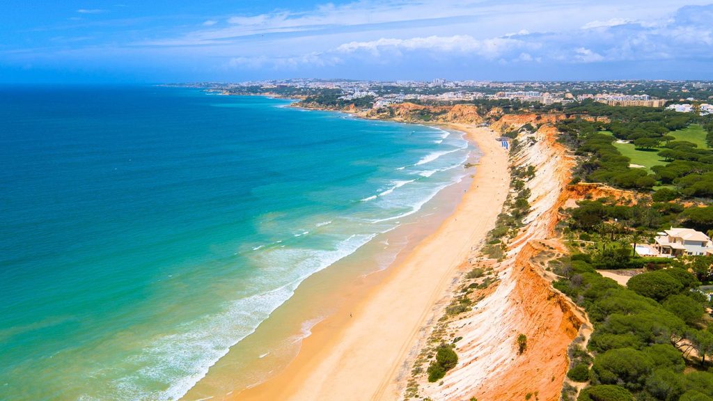 Aerial view of a sandy beach lined with red cliffs and pine trees, meeting the blue ocean under a partly cloudy sky.