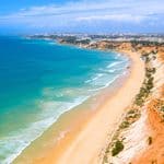 Aerial view of a sandy beach lined with red cliffs and pine trees, meeting the blue ocean under a partly cloudy sky.