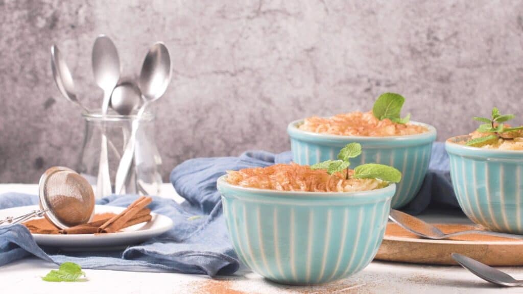 Two teal bowls of rice pudding with cinnamon and mint, next to a plate with a tea strainer and cinnamon sticks, a perfect fall dessert recipe without pumpkin.
