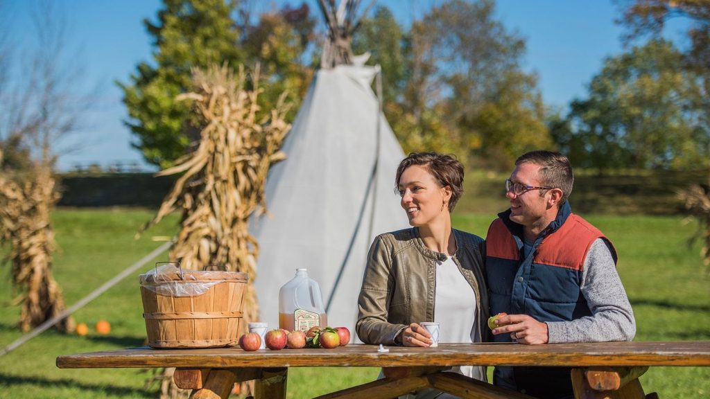 A couple sits at a picnic table with a basket of apples, a jug of cider, and a tepee in the background.