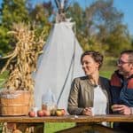 A couple sits at a picnic table with a basket of apples, a jug of cider, and a tepee in the background.
