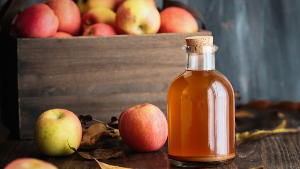 A glass bottle of fresh fall cider with a cork stopper, placed in front of a wooden crate filled with apples and a few loose apples on a table.