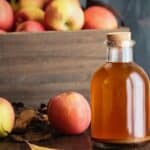A glass bottle of fresh fall cider with a cork stopper, placed in front of a wooden crate filled with apples and a few loose apples on a table.