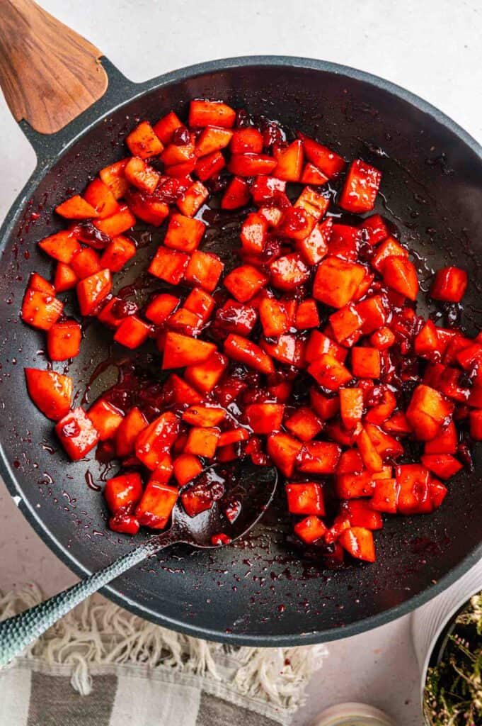 Diced red fruit or vegetables glazed in a dark sauce in a frying pan with a serving spoon.