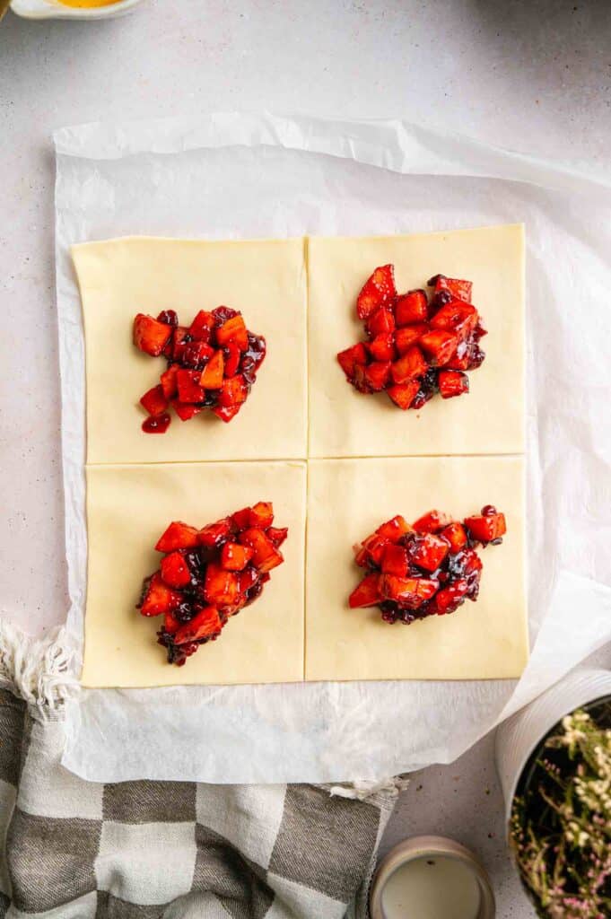 Four square pieces of puff pastry dough on parchment paper, each topped with a portion of cooked strawberry filling.