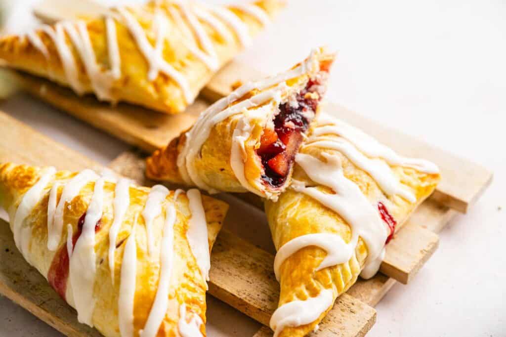 Three triangular fruit turnovers drizzled with white icing on a wooden rack, with one split open to reveal a red berry filling.