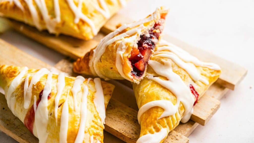 Three fruit-filled pastry turnovers drizzled with white icing, one split open to show a berry filling.