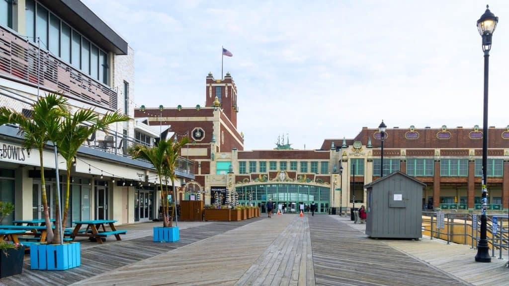 A wooden boardwalk leading toward the Paramount Theatre building in Asbury Park, with palm trees in blue planters and a small ticket booth.