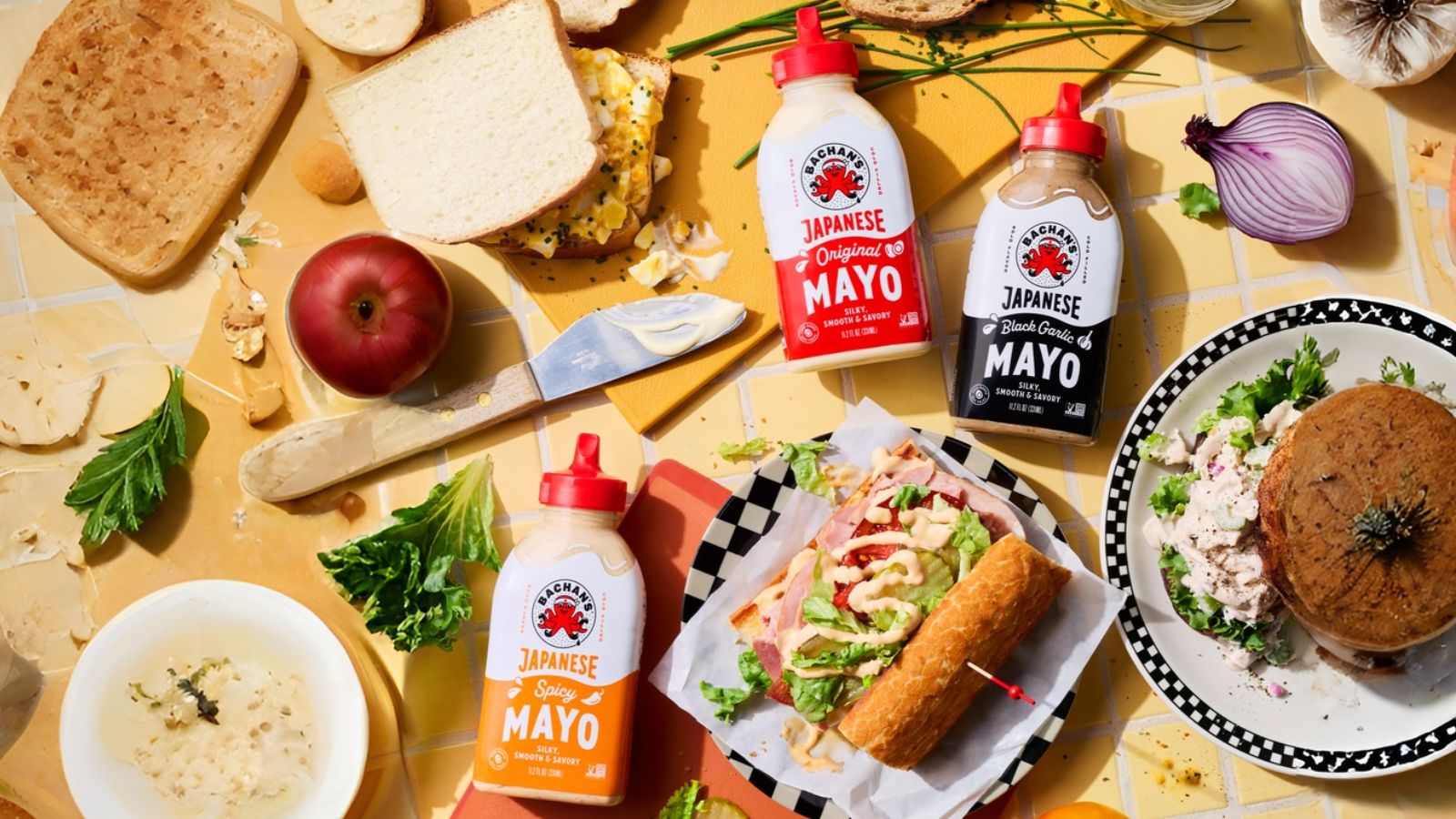 Three squeeze bottles of Bachan&rsquo;s Japanese mayo&mdash;original, black garlic, and spicy&mdash;surrounded by sandwiches, salad, and loose ingredients on a yellow tiled surface.