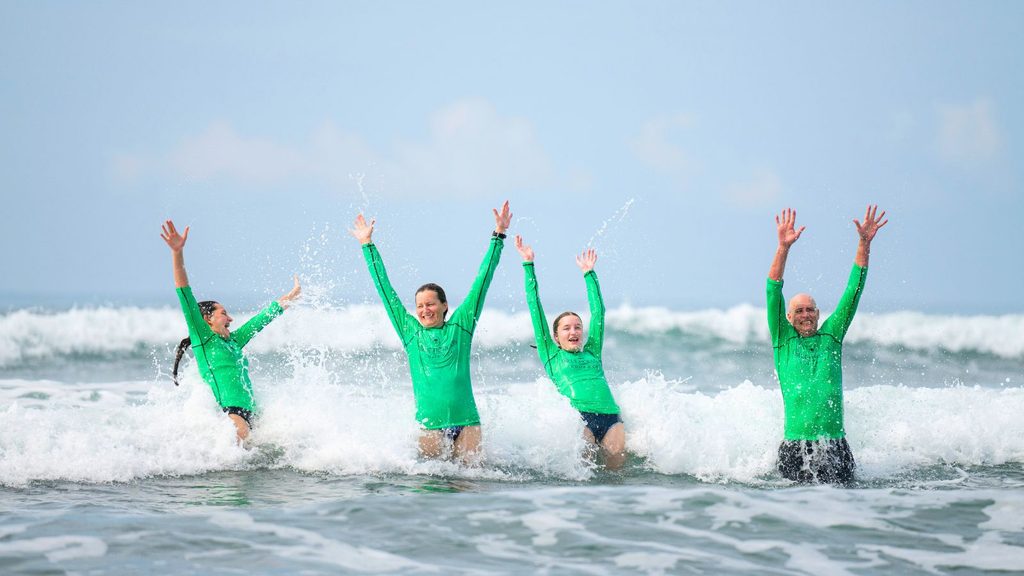 Four people in matching green long-sleeved shirts stand in ocean waves with their arms raised.
