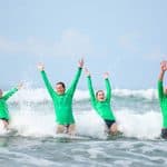 Four people in matching green long-sleeved shirts stand in ocean waves with their arms raised.