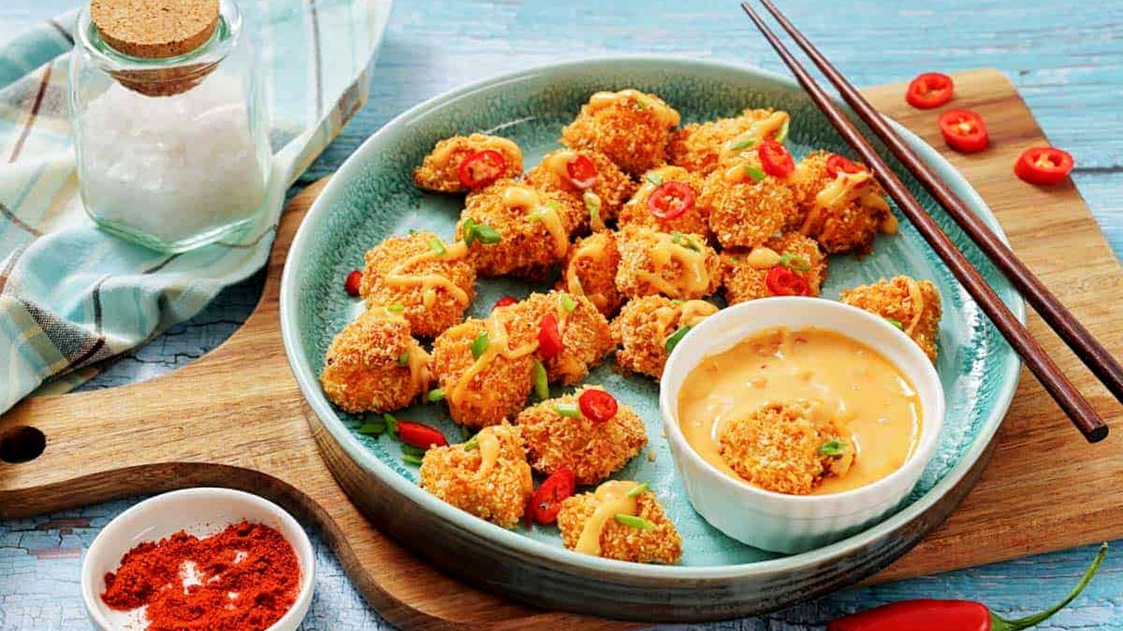 A plate of bang bang chicken bites garnished with sliced red chili and green onions, served with a dipping sauce.