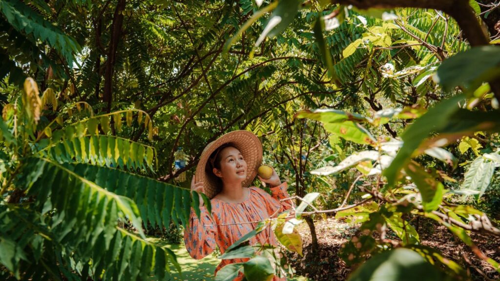 A woman wearing a wide-brimmed straw hat and an orange patterned top holds a yellow fruit while standing in a leafy garden, perfect for closed-door travel.