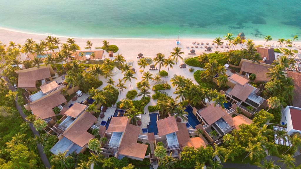 Aerial view of a coastal resort featuring thatched-roof villas, swimming pools, and palm trees along a sandy beach.