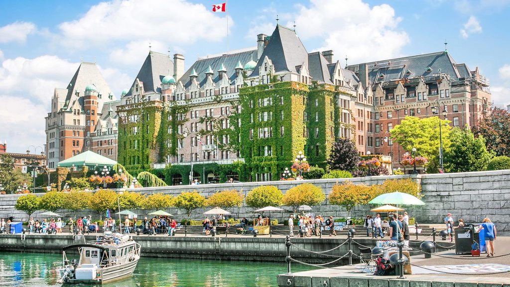 The ivy-covered Fairmont Empress overlooks the Inner Harbour in Victoria, BC, near local bookstores, as people walk along the stone quay in the foreground.