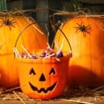 An orange jack-o’-lantern trick-or-treat bucket filled with lollipops sits in front of two large pumpkins decorated with plastic spiders on straw.