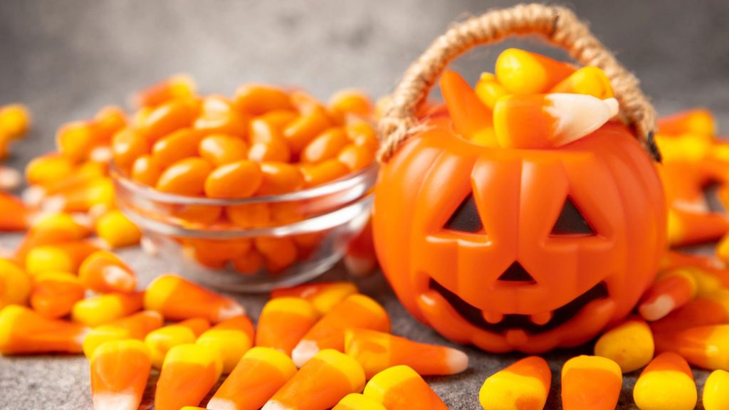 A plastic jack-o&rsquo;-lantern bucket filled with candy corn for National Candy Corn Day, beside a glass bowl of orange jelly beans, surrounded by loose candy corn.