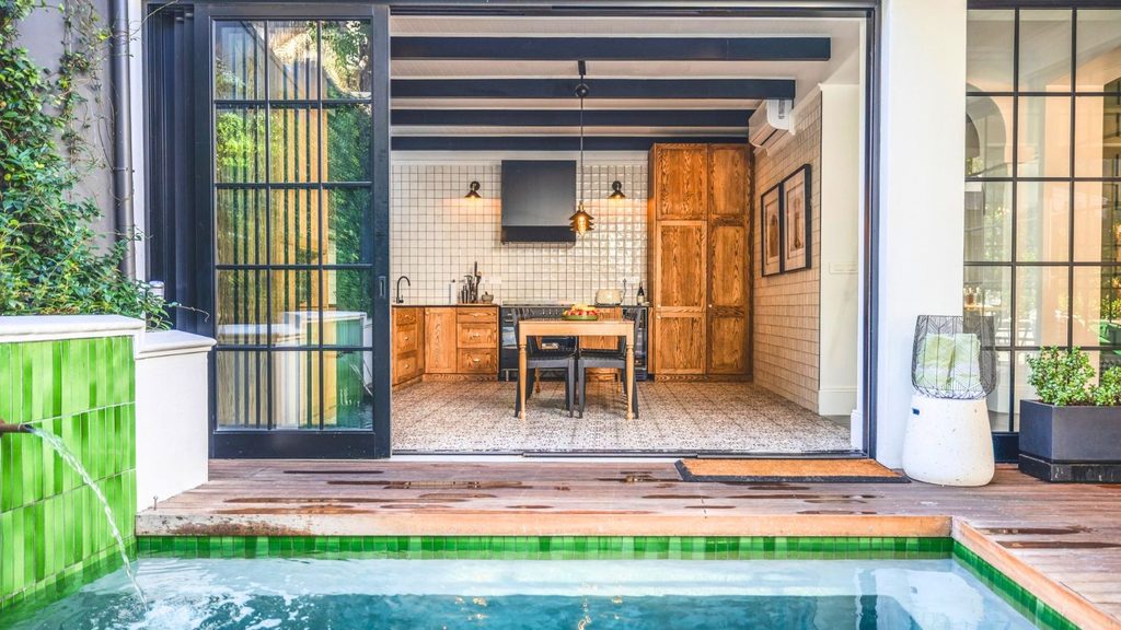 A kitchen with wooden cabinets and a dining table, seen from a patio with a swimming pool.