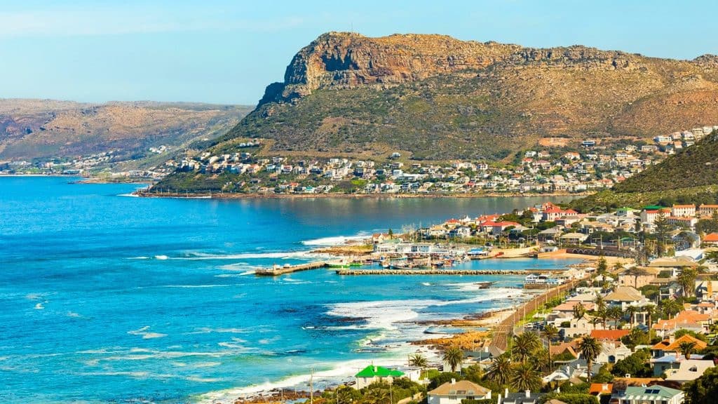 A view of the coastal town of Kalk Bay with a harbor, turquoise water, and a mountain rising in the background.