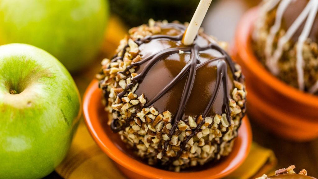 Caramel apple dipped in nuts and drizzled with chocolate, sitting in an orange bowl next to a fresh green apple.