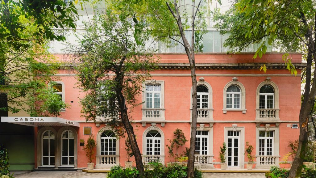 The pink facade of a Casona building with arched windows, stone balconies, and a white entrance canopy, surrounded by trees.