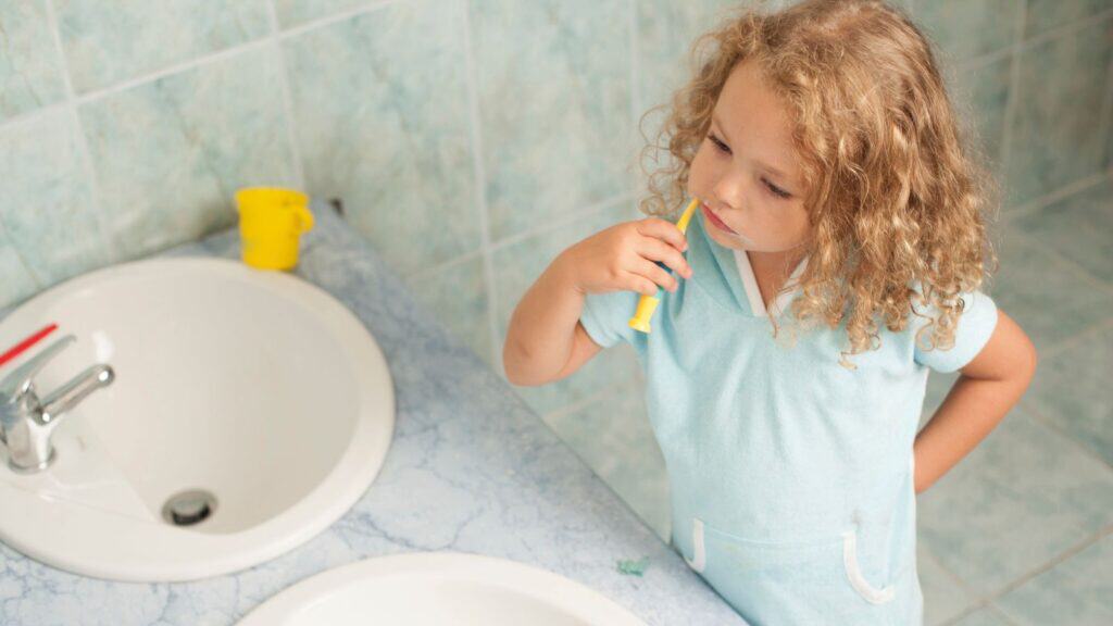A young child with curly hair brushes their teeth at a bathroom sink, perhaps prepping for Halloween tricks.