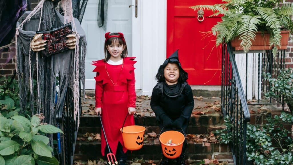 Two children in Halloween costumes hold pumpkin candy buckets on a front porch, highlighting the holiday excitement and the significant amount Americans spend on Halloween.