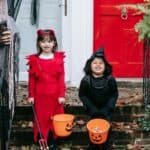 Two children in Halloween costumes hold pumpkin candy buckets on a front porch, highlighting the holiday excitement and the significant amount Americans spend on Halloween.