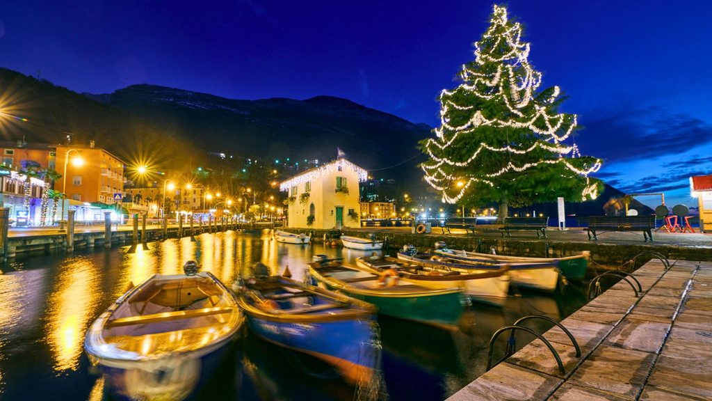 Small boats moored in a harbor at dusk, featuring a large, illuminated Christmas tree on the shore.