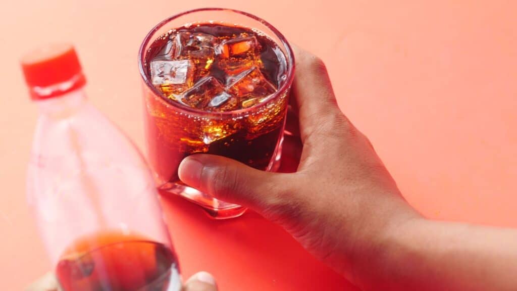 A hand holds a glass of ice-filled dark soda, a hard-to-find regional soda, next to a partially visible plastic bottle.