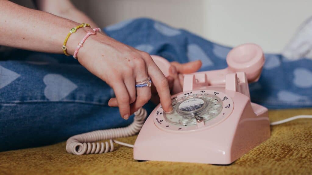 A hand dials a pink rotary telephone, reminiscent of the 90s kids era.