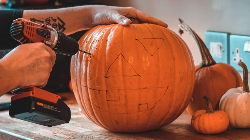 A person using a cordless drill as one of their pumpkin carving techniques on a jack-o&rsquo;-lantern with a face drawn in marker.