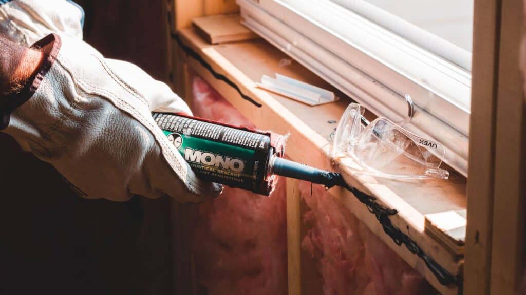 A gloved hand applies Mono acoustic sealant to a window frame to weatherproof the home before a cold snap.