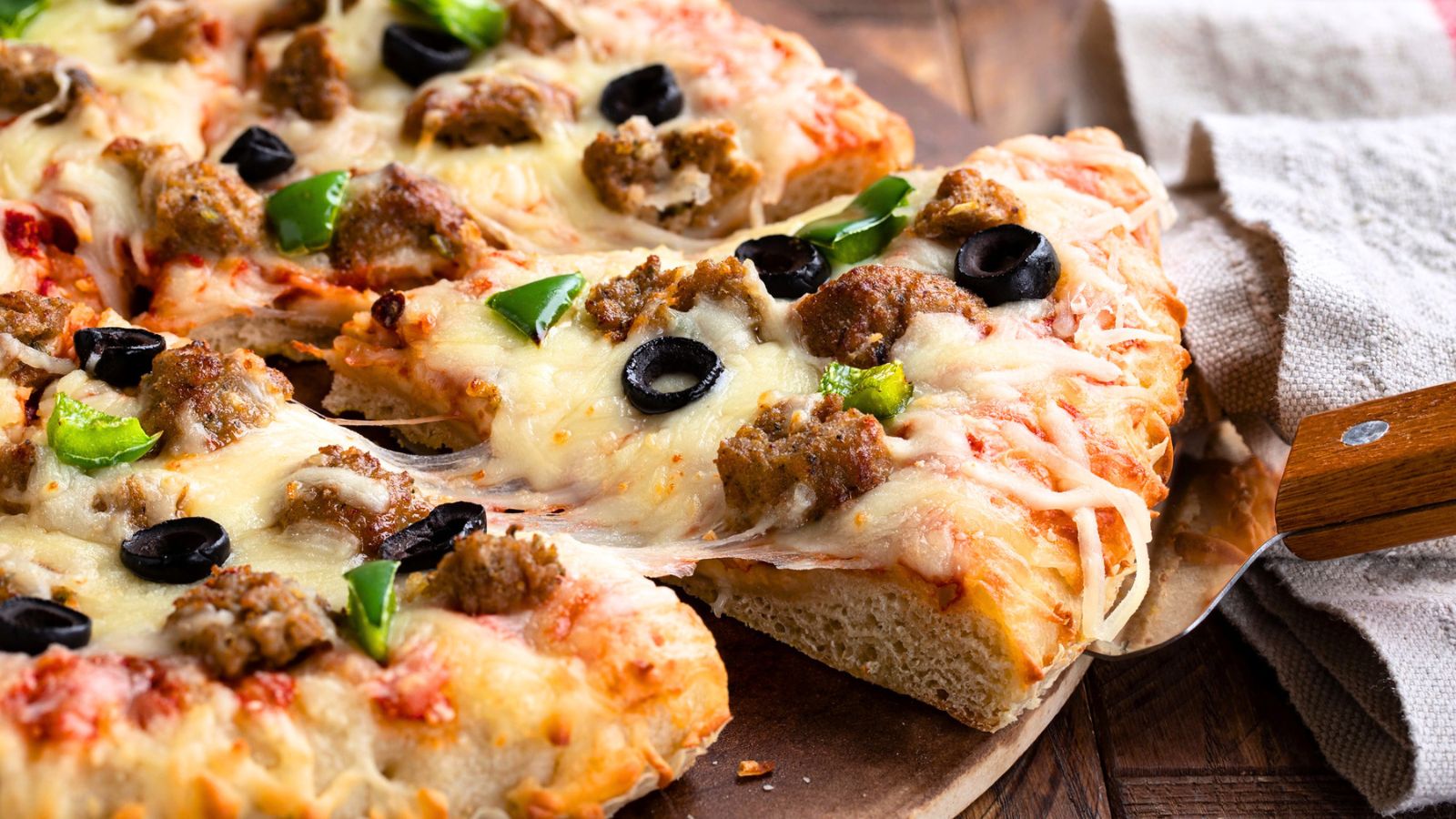 A slice of pizza topped with sausage, black olives, and green peppers being lifted from a wooden board.