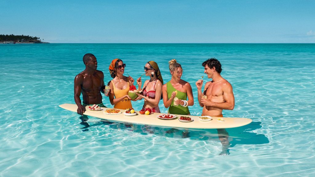 Five people in swimwear stand in clear ocean water around a surfboard acting as a table, eating fruit and drinking from coconuts.