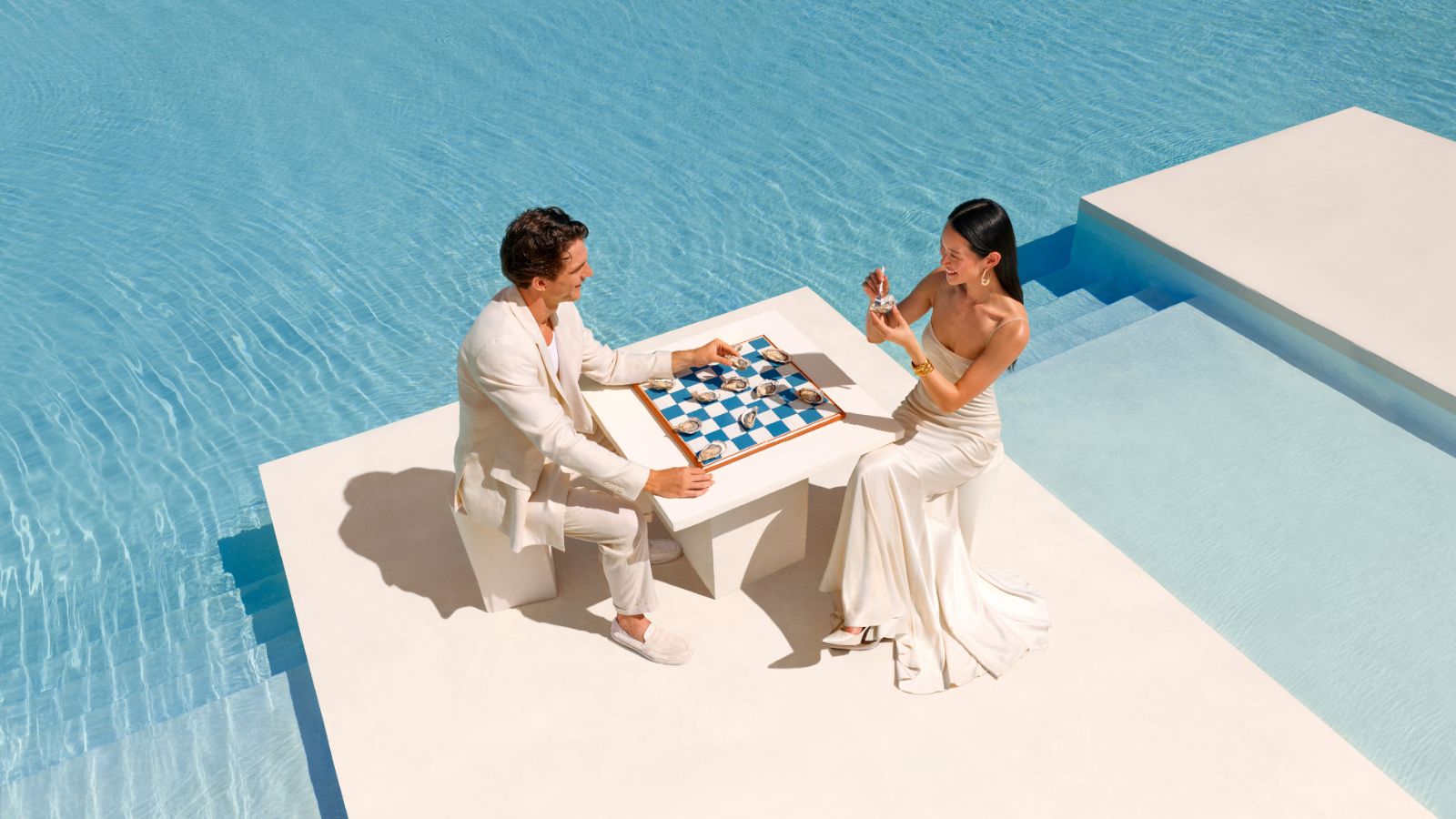 A man and woman in formal attire play a game on a blue and white checkered board at a Club Med, set on a small table on a white platform surrounded by a swimming pool.