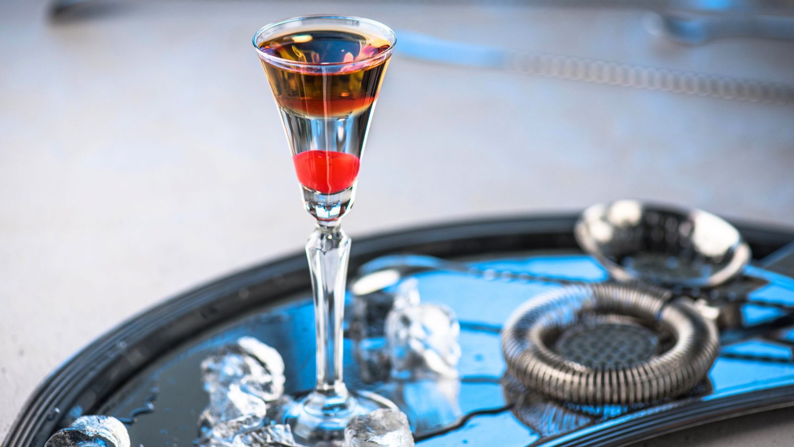 A stemmed cocktail glass containing a layered, two-tone tiny cocktail with a maraschino cherry at the bottom, resting on a dark tray with ice and a strainer.