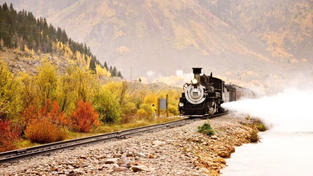 A steam train travels along tracks beside a river in a mountainous landscape, offering scenic train rides through vibrant fall foliage.