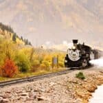 A steam train travels along tracks beside a river in a mountainous landscape, offering scenic train rides through vibrant fall foliage.