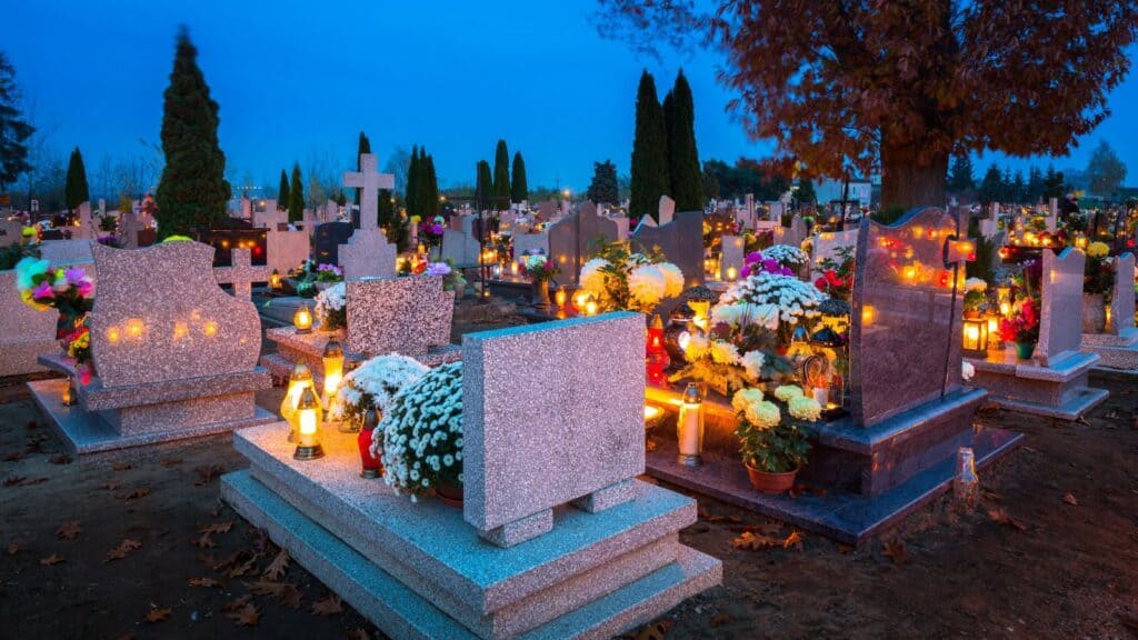 A cemetery at twilight during Halloween-like traditions, with many gravestones decorated with potted flowers and glowing votive candles.
