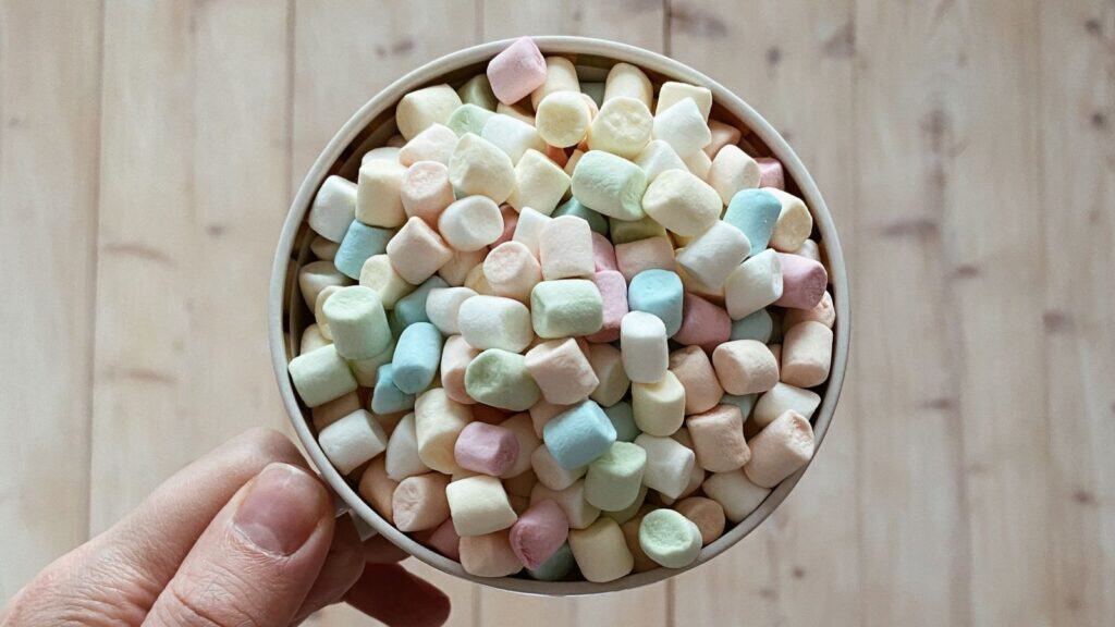 A hand holding a mug filled with miniature pastel-colored marshmallows suitable for caramel apple toppings.