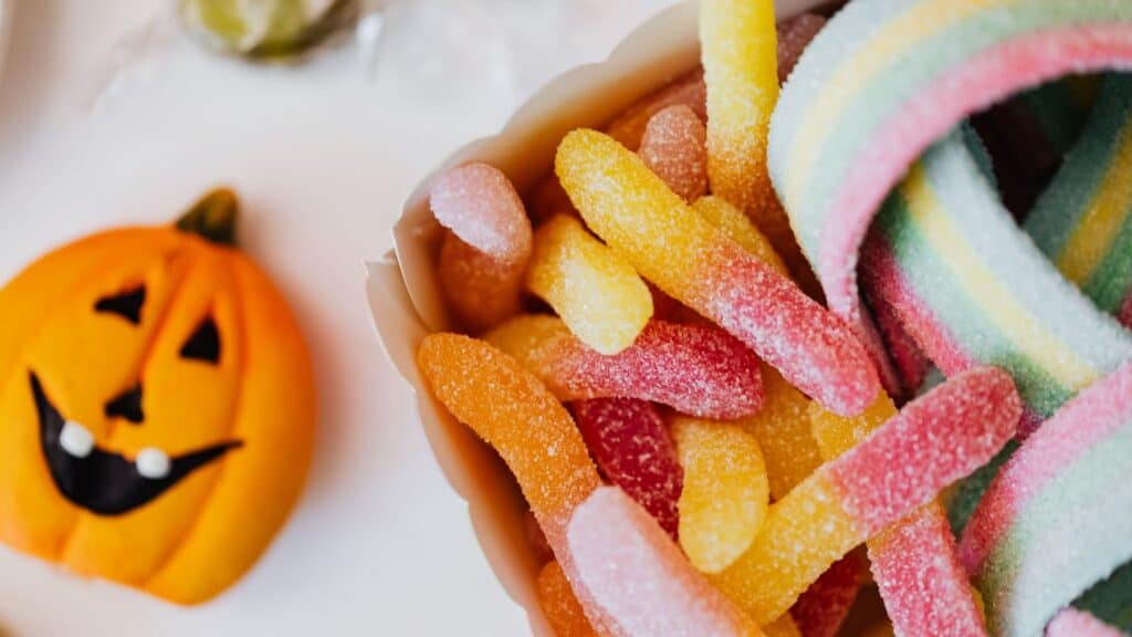 A paper bowl filled with colorful sugar-coated gummy worms and an orange pumpkin-shaped treat, perfect for those looking to make Halloween candy last longer.